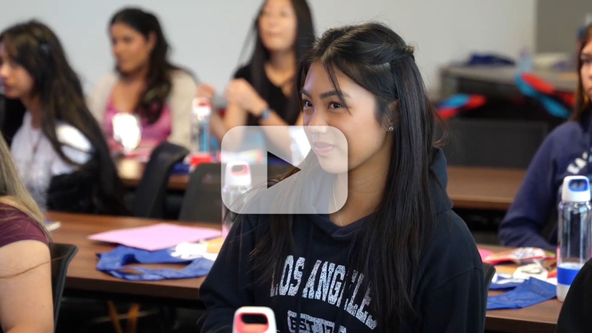 A young student sits amongst her FNL peers as they receive a presentation, with video play button.