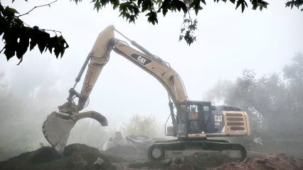San Bernardino County Public Works excavator moving soil on a foggy morning, surrounded by trees.