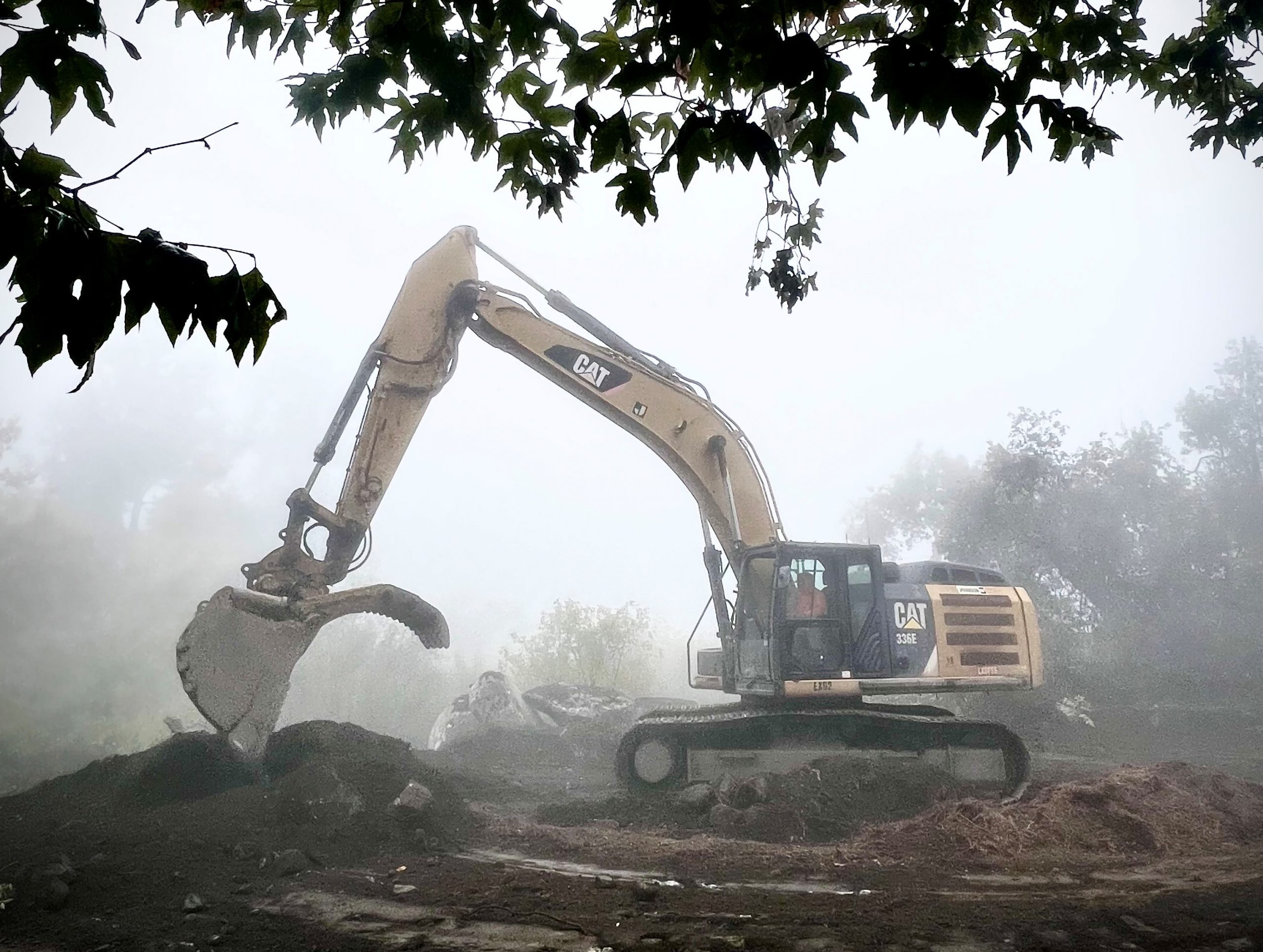San Bernardino County Public Works excavator moving soil on a foggy morning, surrounded by trees.