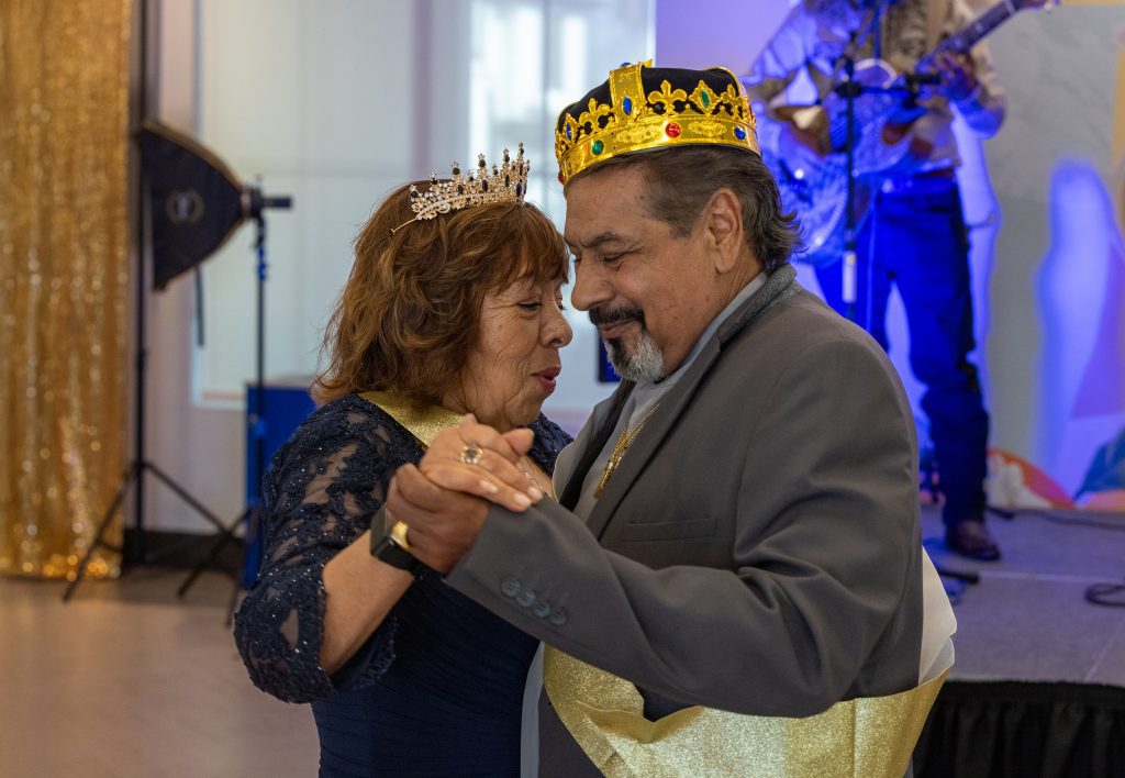 An elderly couple wearing crowns and sashes dance closely, smiling warmly at a celebration with a musician playing in the background.