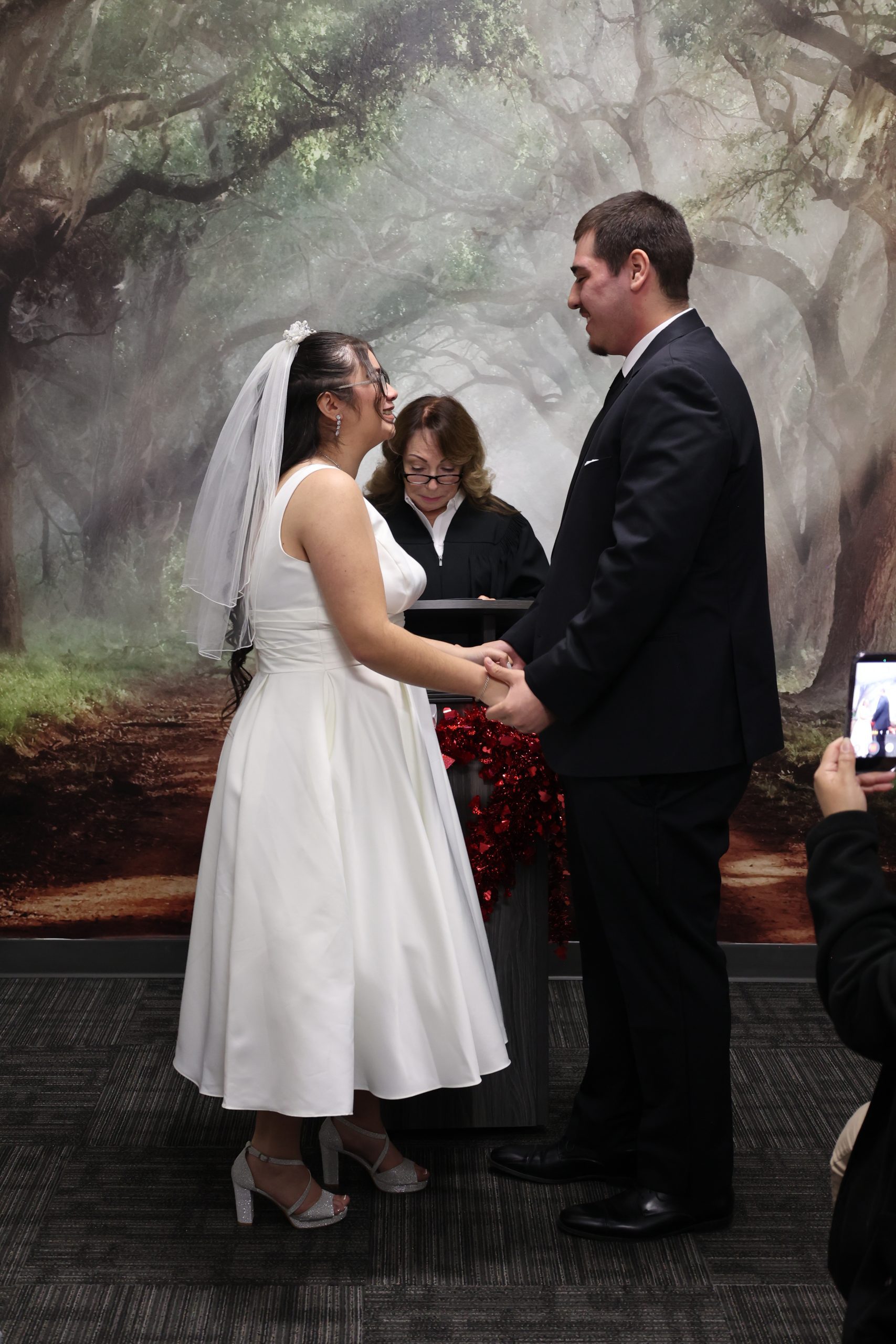 Bride and groom hold hands and smile during a wedding ceremony as an officiant reads from a podium before a forest backdrop.