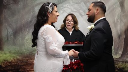 Bride and groom hold hands and smile during a wedding ceremony as an officiant stands between them against a forest backdrop.