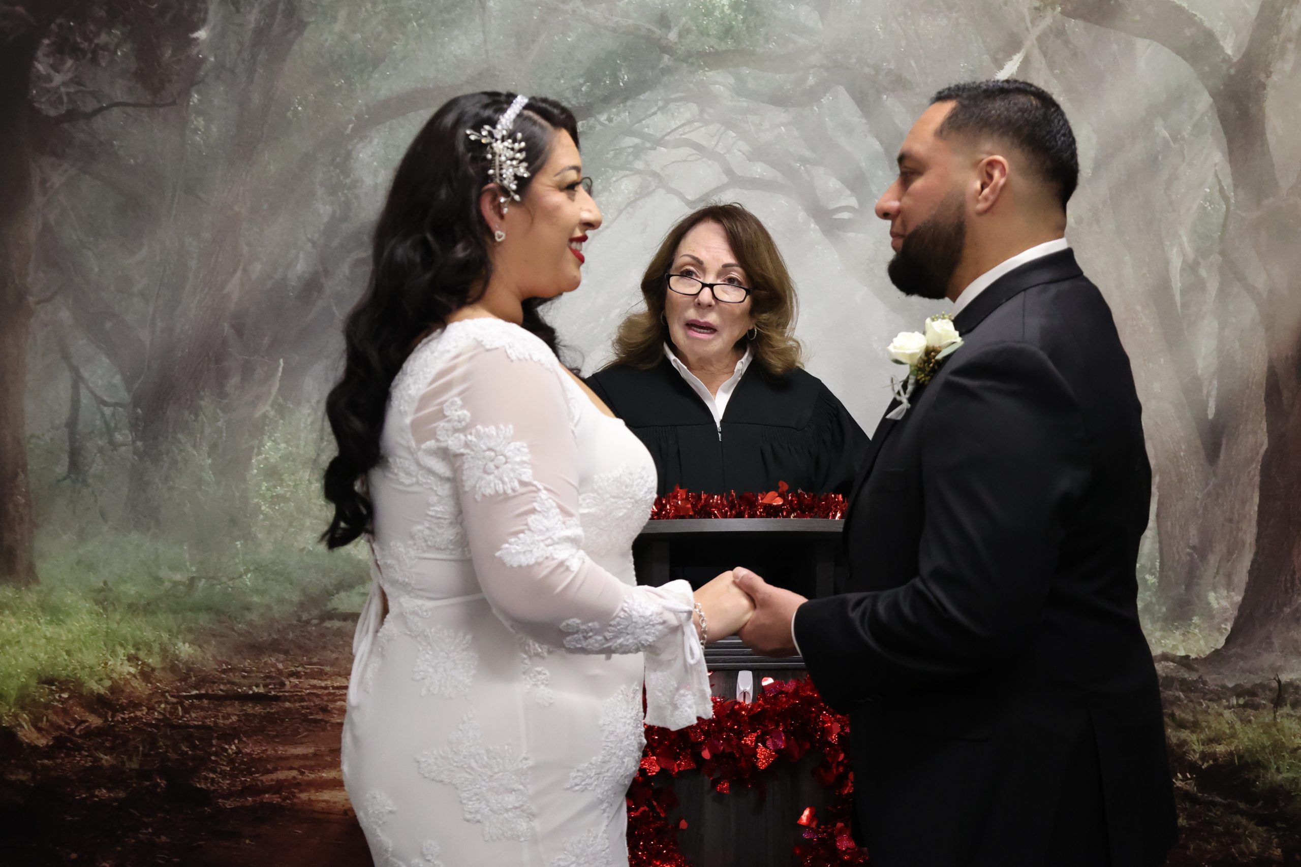 Bride and groom hold hands and smile during a wedding ceremony as an officiant stands between them against a forest backdrop.