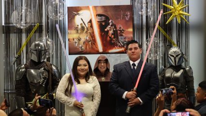 A couple in wedding attire holds lightsabers, surrounded by people in sci-fi costumes. A "Star Wars" poster and festive decorations are in the background.