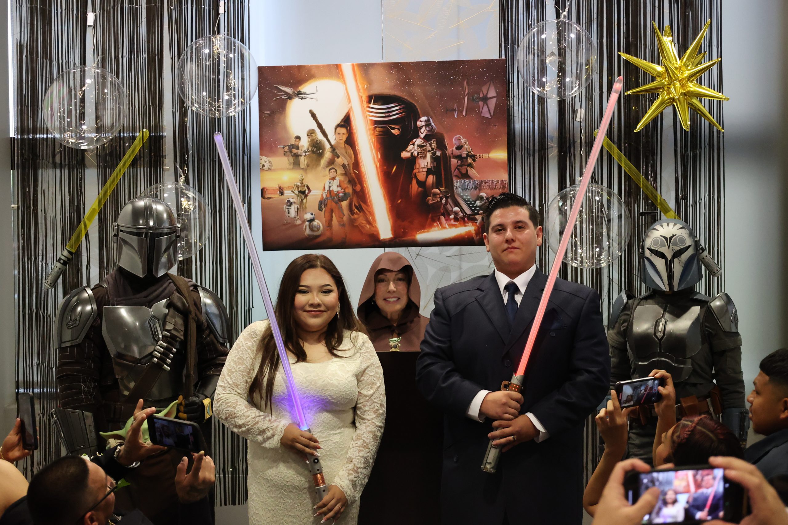 A couple in wedding attire holds lightsabers, surrounded by people in sci-fi costumes. A "Star Wars" poster and festive decorations are in the background.