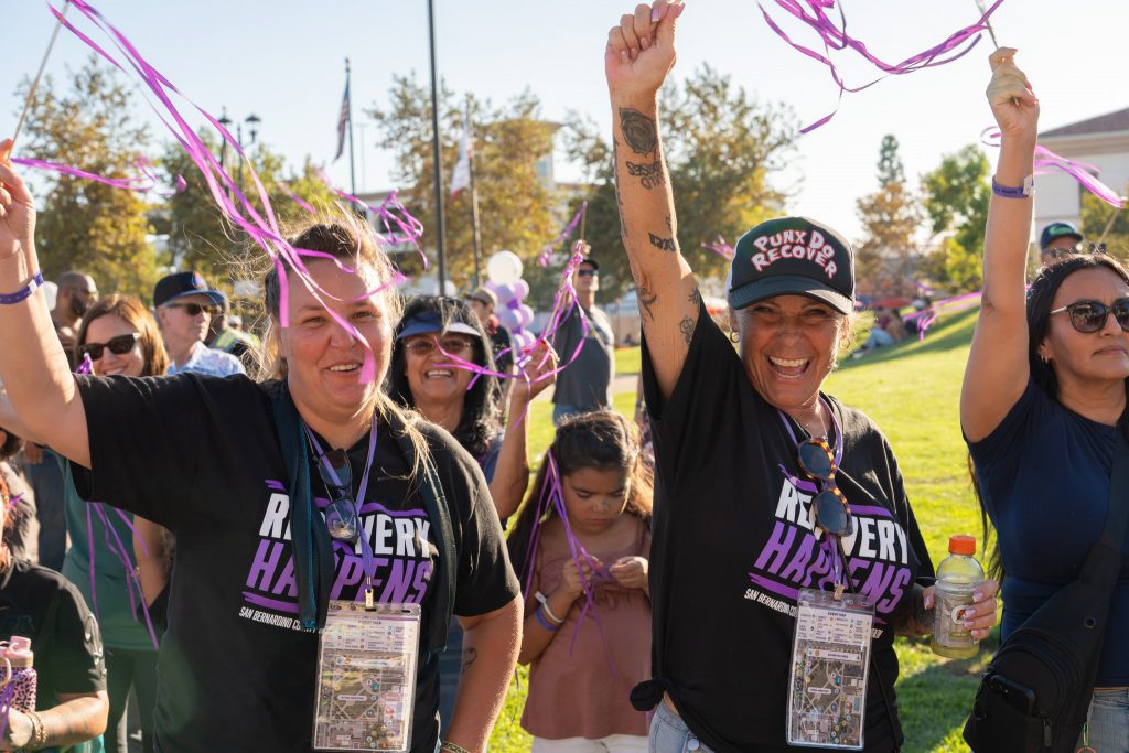 Recovery Happens attendees celebrate outdoors, smiling and waving purple streamers during a community event in San Bernardino County.