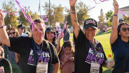Recovery Happens attendees celebrate outdoors, smiling and waving purple streamers during a community event in San Bernardino County.