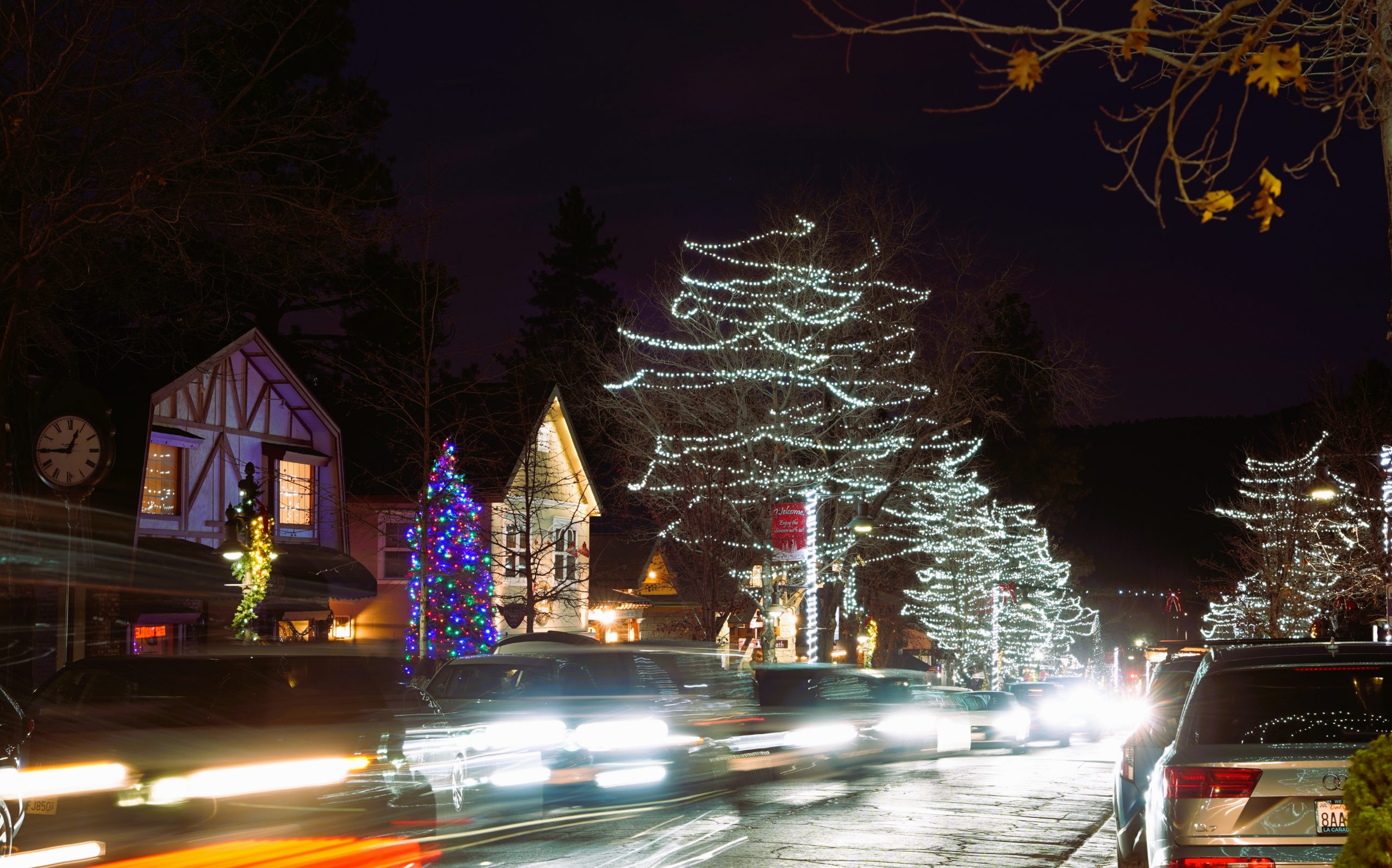 Nighttime view of cars driving down a street with bright headlights beaming, along a row of homes and trees with holiday lights.