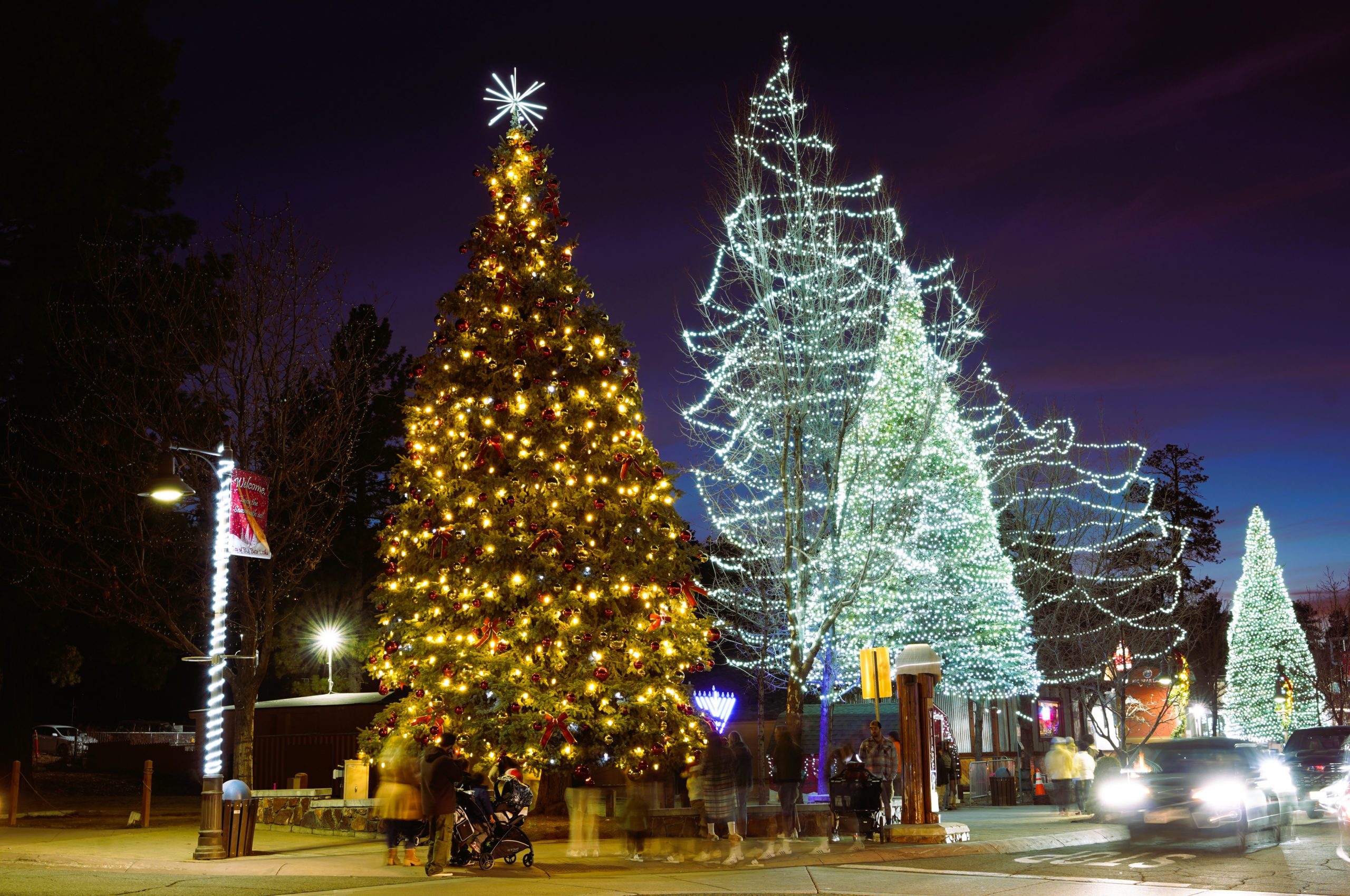 Nighttime view of a busy street in Big Bear Lake, with holiday decorations, including a several trees decorated with Christmas lights.
