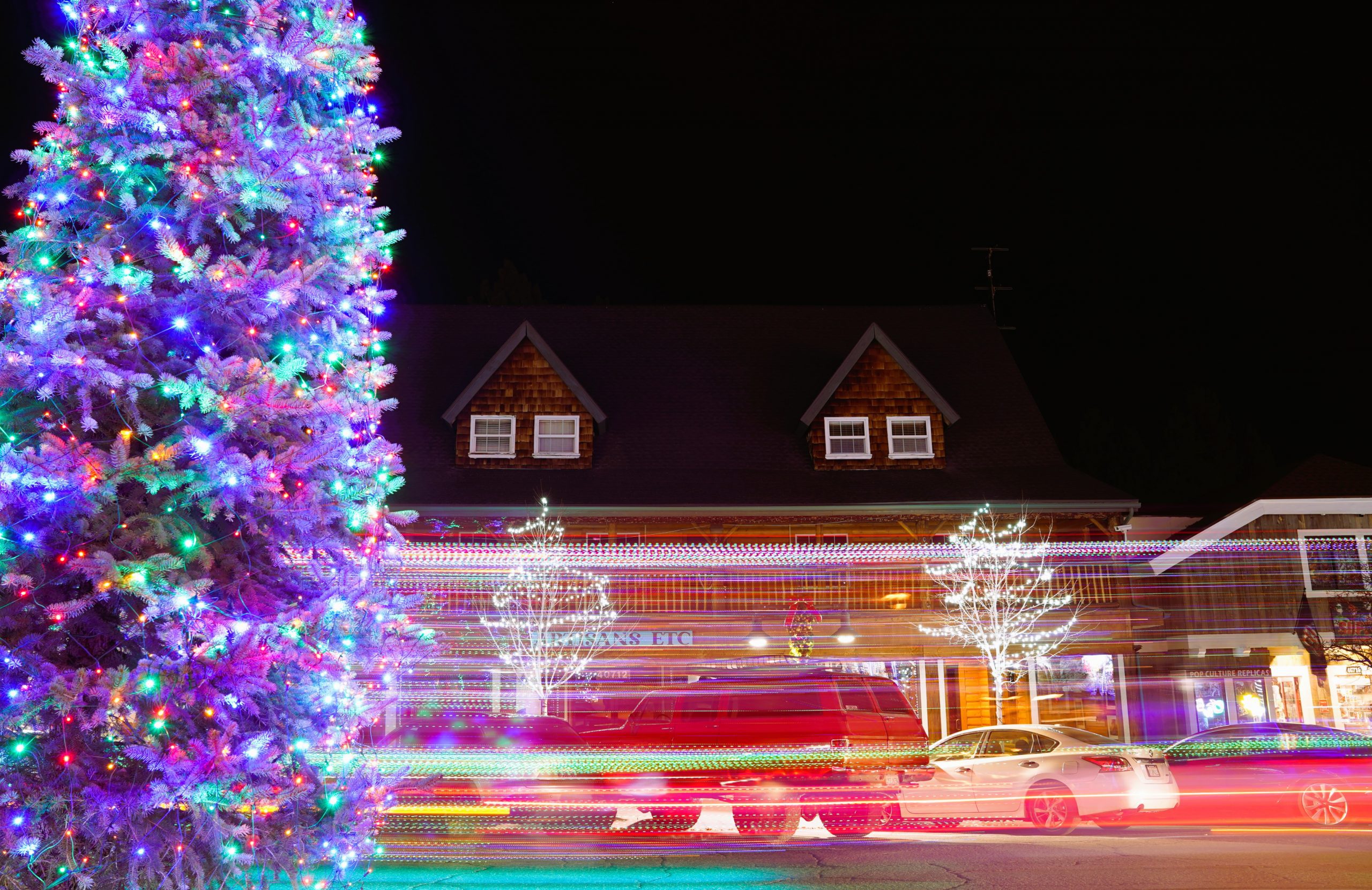 Several trees covered in holiday lights with blurred cars passing by several buildings.