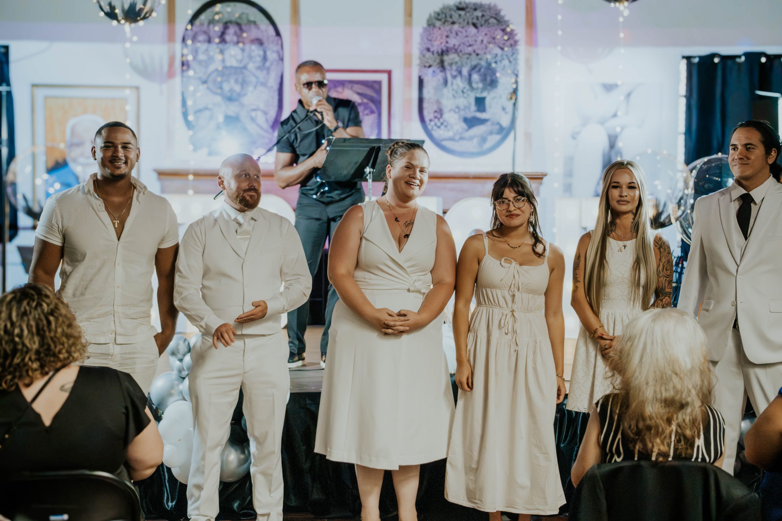 Six people stands at the front of a room decorated for an event, with onlookers seated around them and balloons in the background. An artist performs in the background. Everyone is dressed in formal white attire.
