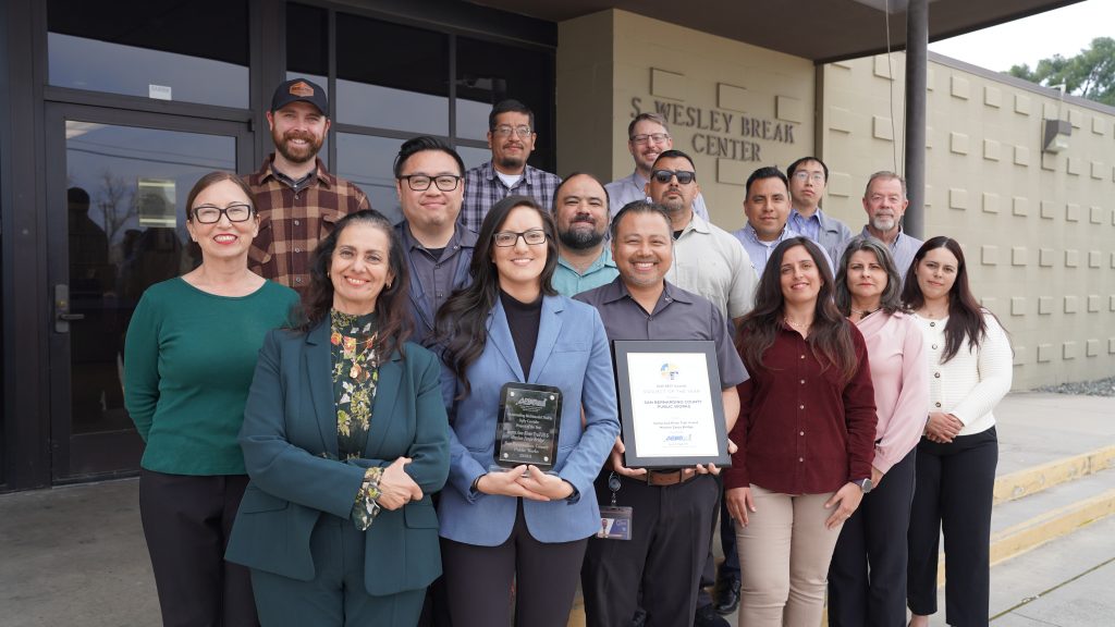 Group of Public Works employees standing outside a building, smiling as two people hold plaques recognizing a completed project.