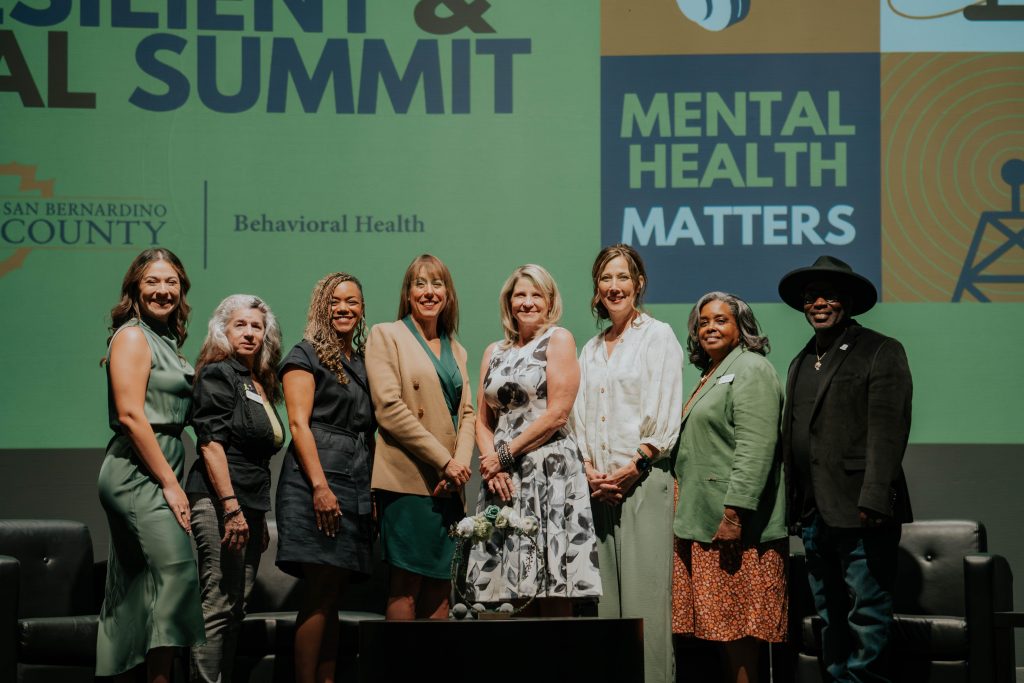 Eight individuals stand together on stage at the San Bernardino County Mental Health Summit, smiling in front of a green backdrop with event signage.