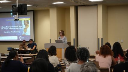 A person is speaking at a podium in a conference room, with an audience seated at tables. A presentation slide about "Shaken Baby Syndrome and Infant Safe Sleep" by San Bernardino County Department of Public Health is projected on a screen.