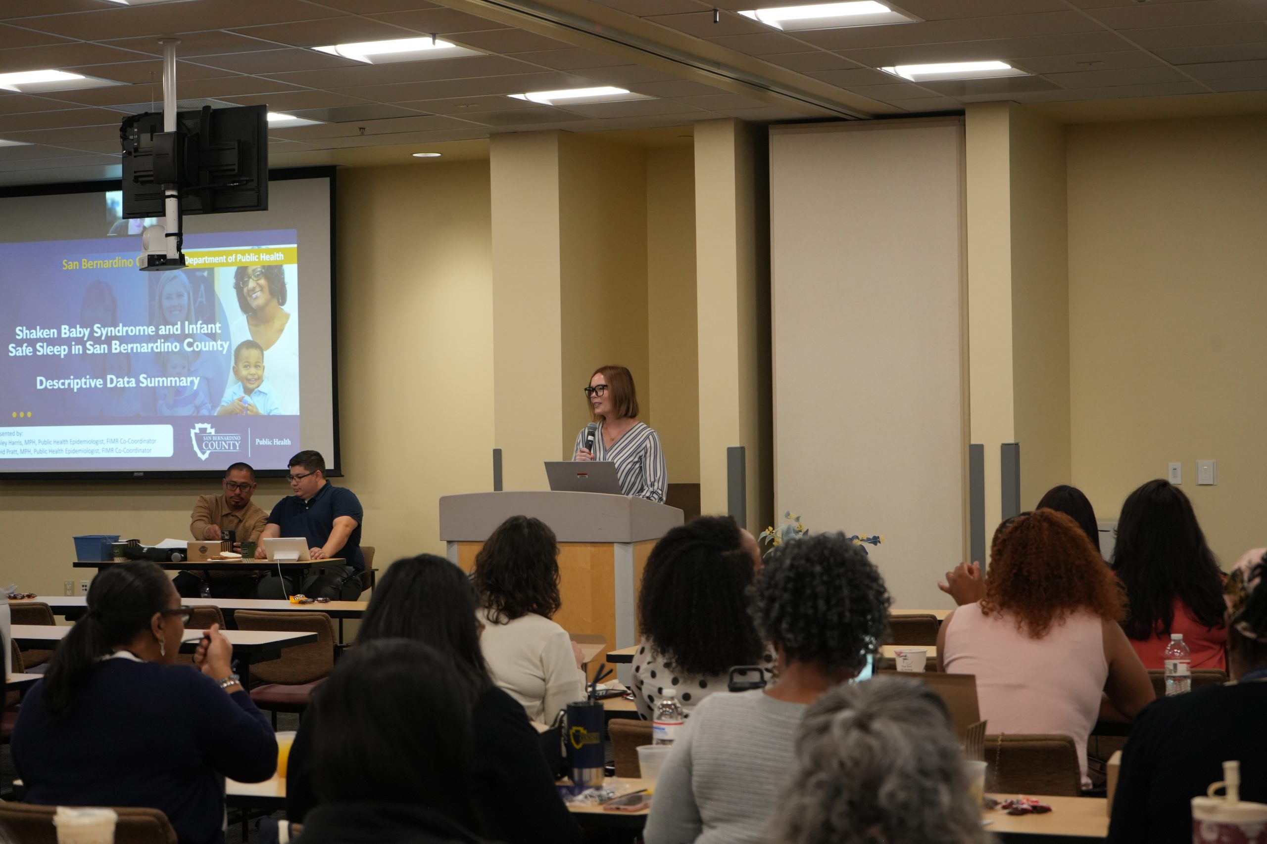 A person is speaking at a podium in a conference room, with an audience seated at tables. A presentation slide about "Shaken Baby Syndrome and Infant Safe Sleep" by San Bernardino County Department of Public Health is projected on a screen.