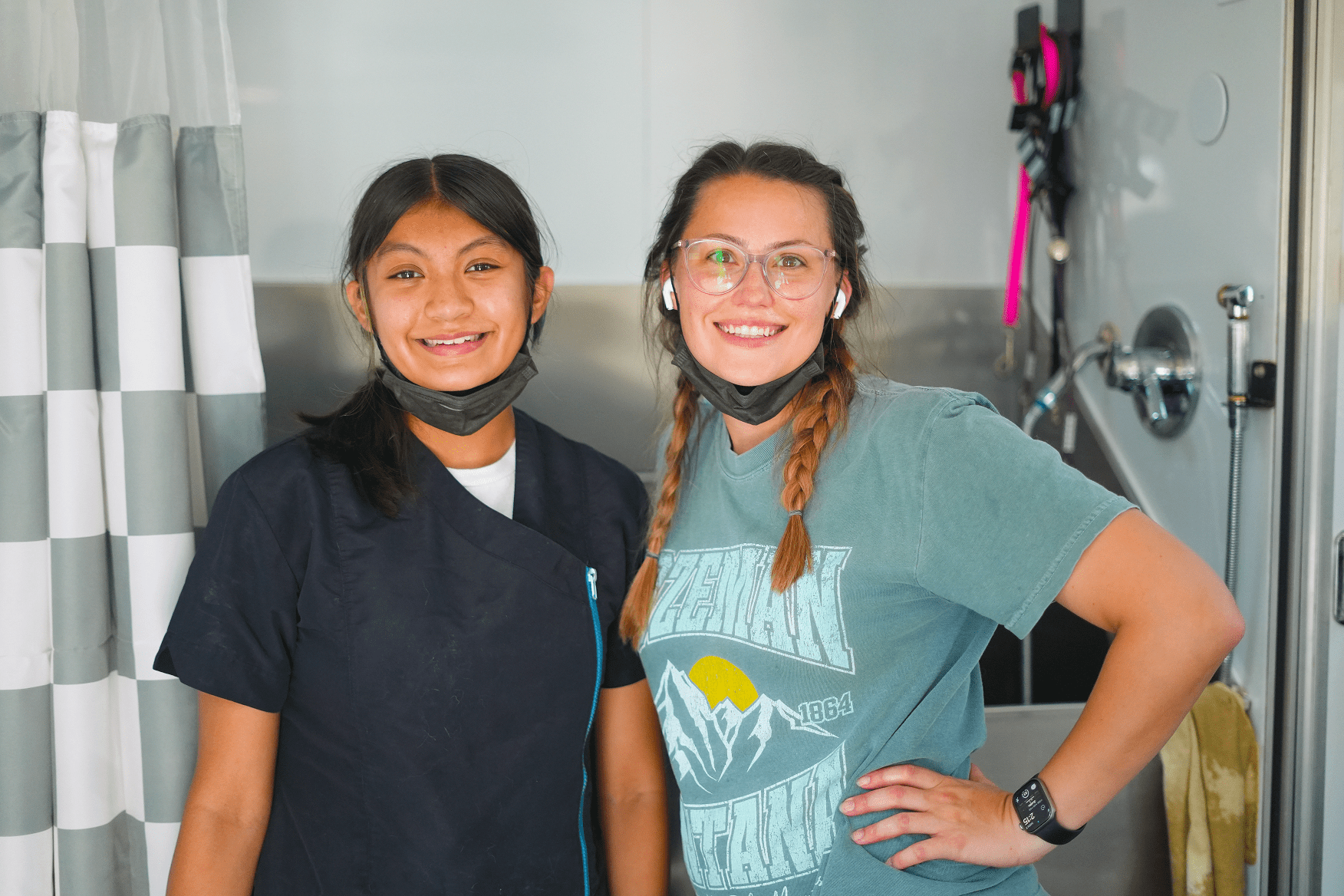 Two volunteer groomers standing inside a mobile grooming unit, wearing grooming attire and face masks, ready to assist shelter pets.