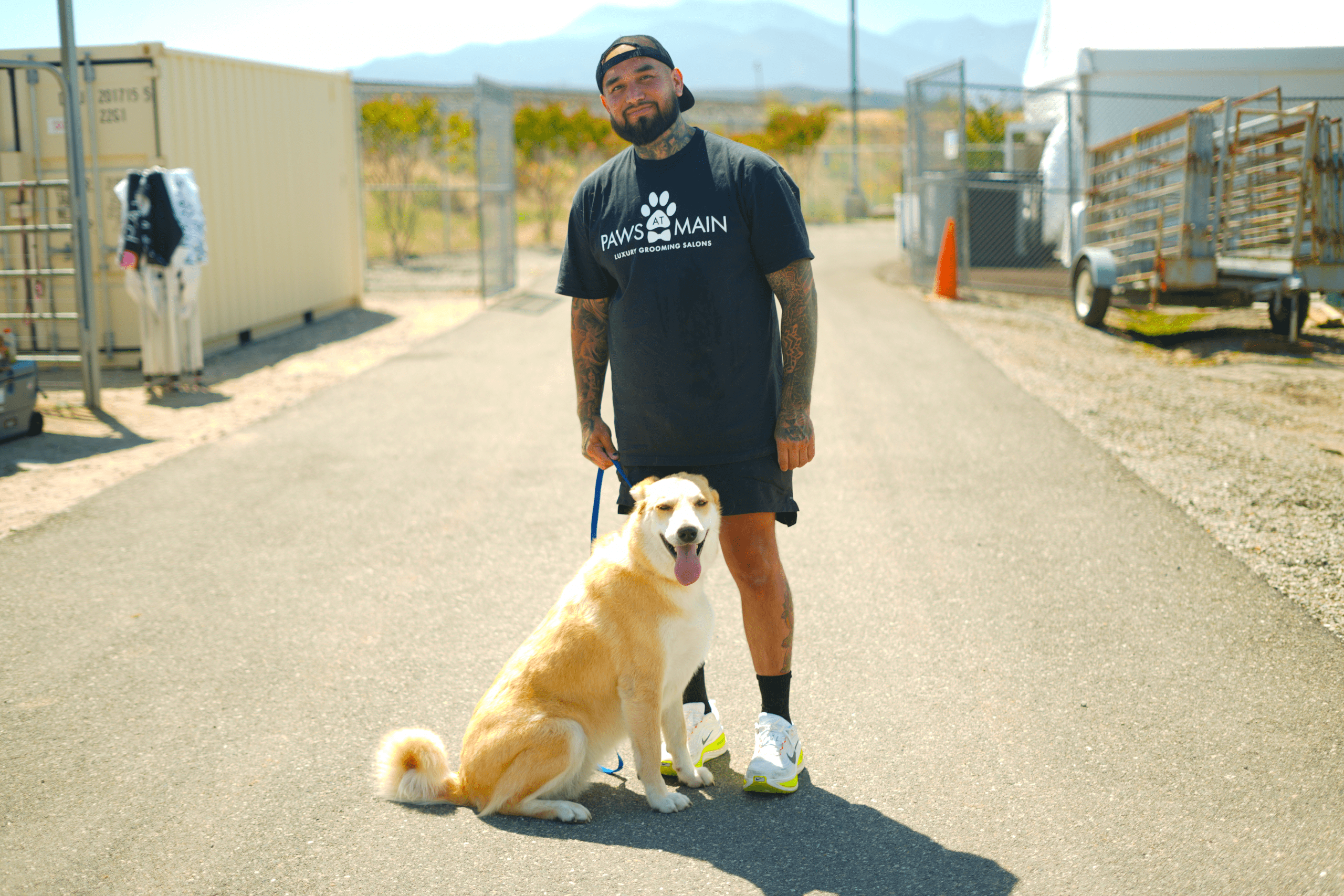 Paws at Main staff standing on an outdoor walkway holding the leash of a dog sitting at their feet, with animal shelter buildings in the background.