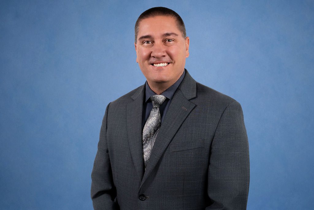 Official portrait of Josh Dugas, Director of the San Bernardino County Behavioral Health Department, in a gray suit against a blue background.