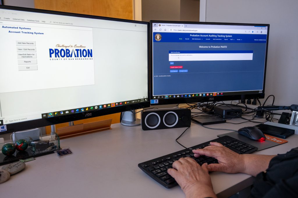 Hands typing on a keyboard with two monitors displaying Probation Account Auditing Tracking System.
