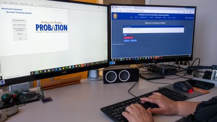 Hands typing on a keyboard with two monitors displaying Probation Account Auditing Tracking System.