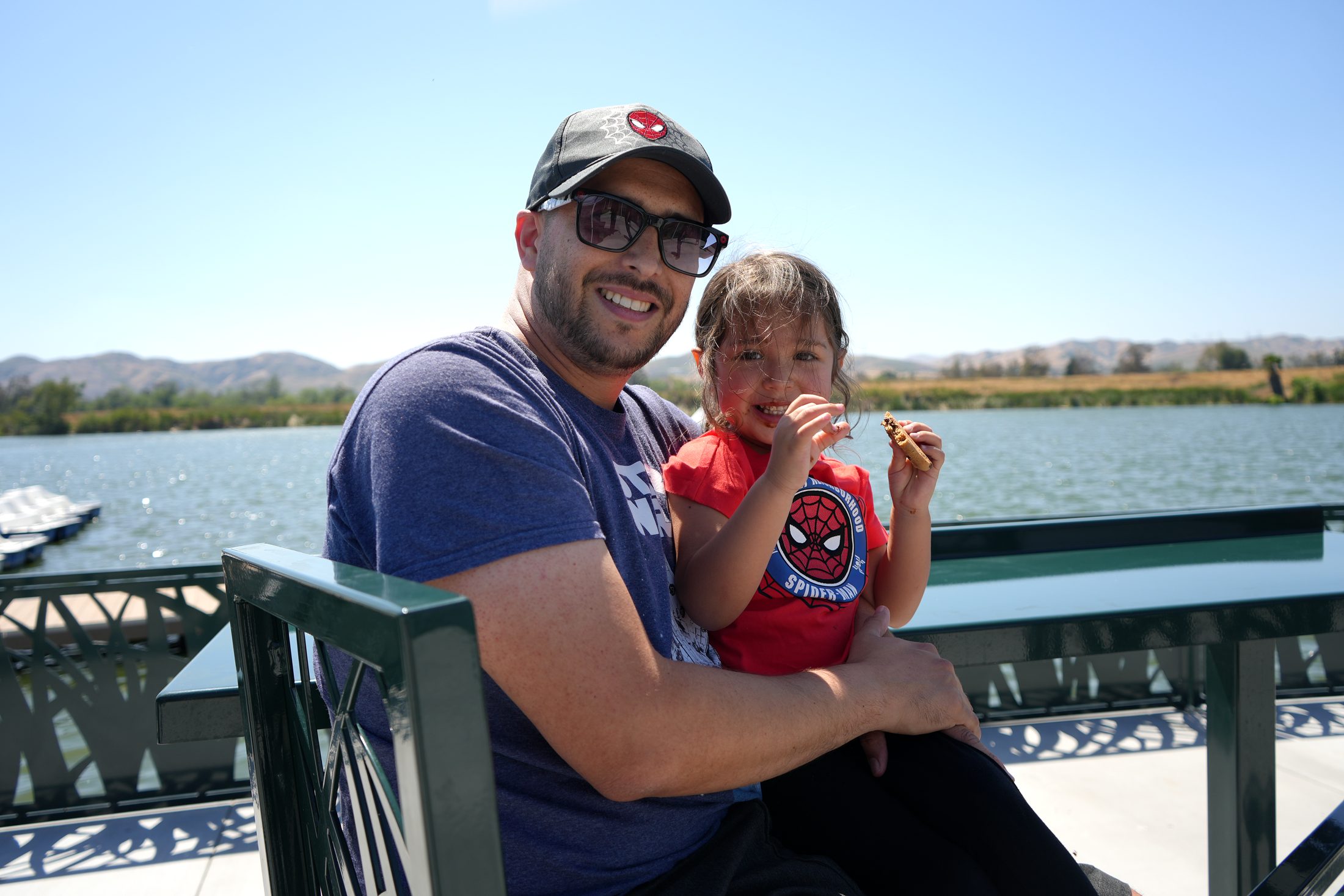 A man holds a little girl in his lap while she eats a chocolate chip cookie with a lake behind them.