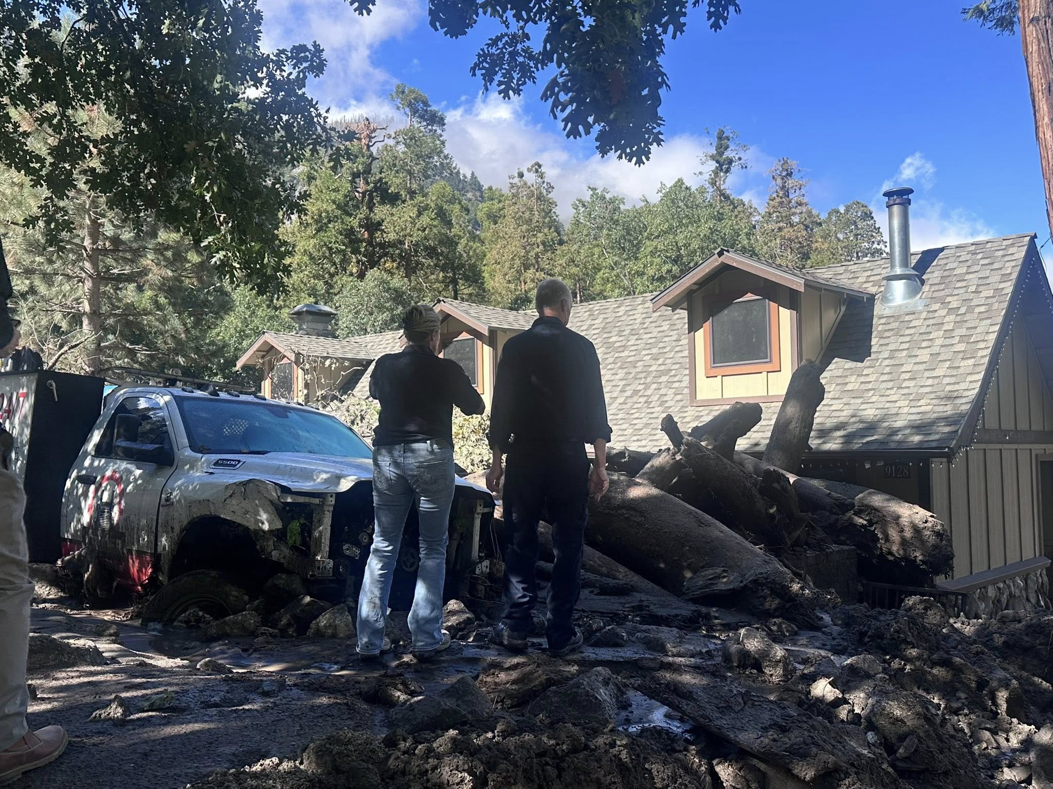 Two people stand near a mud-covered truck and fallen trees in front of a damaged house after a mudslide in a forested area.