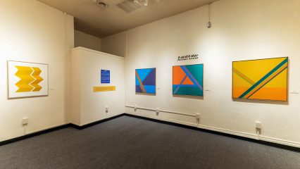 An art exhibition room displaying a series of abstract paintings set against a white wall.