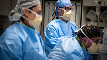 Healthcare professionals in an operating room, all wearing blue surgical gowns, masks and eyewear.