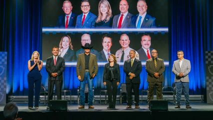Seven individuals standing on a stage wearing formal and casual attire. Behind them is a large screen displaying ten portraits.