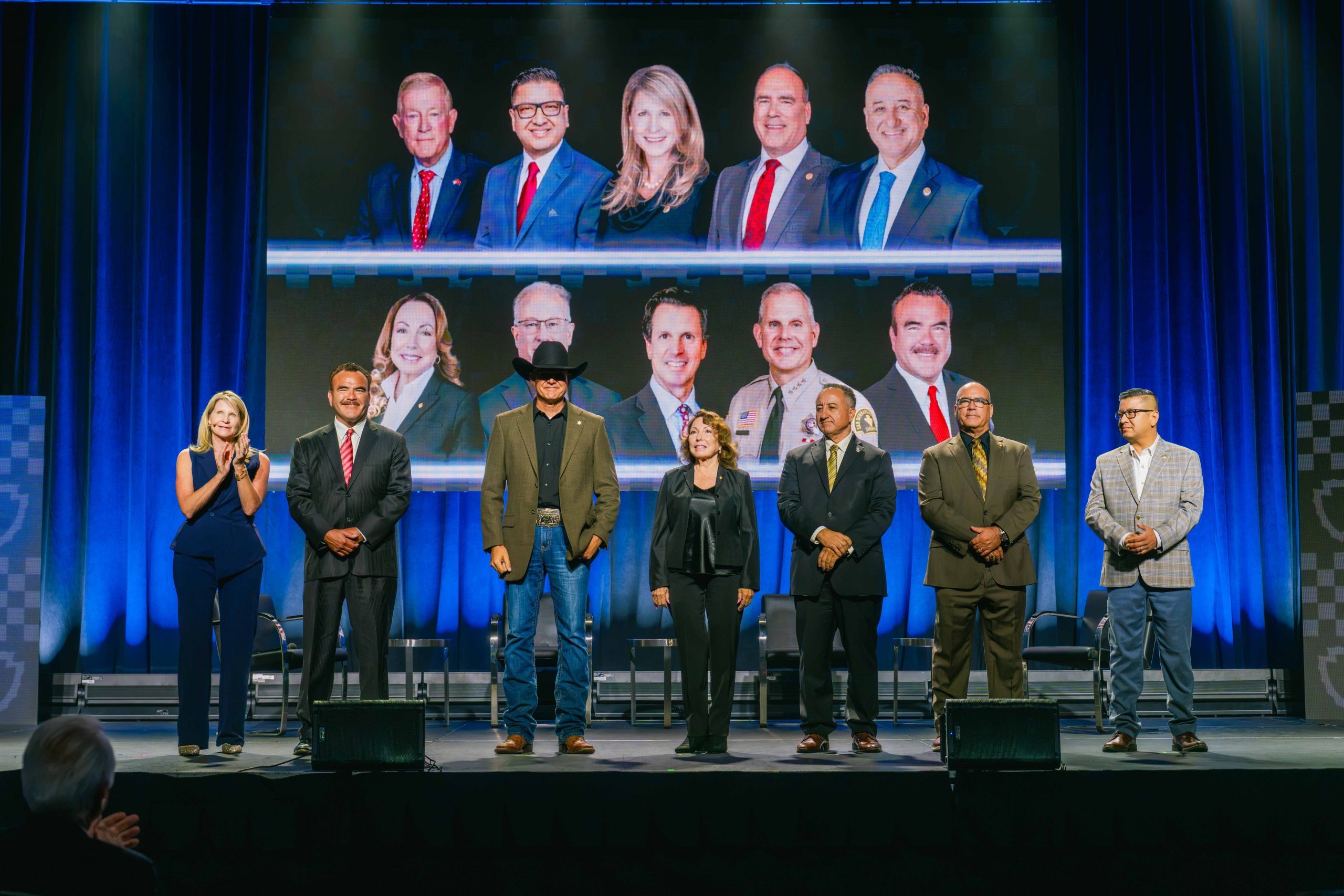 Seven individuals standing on a stage wearing formal and casual attire. Behind them is a large screen displaying ten portraits.