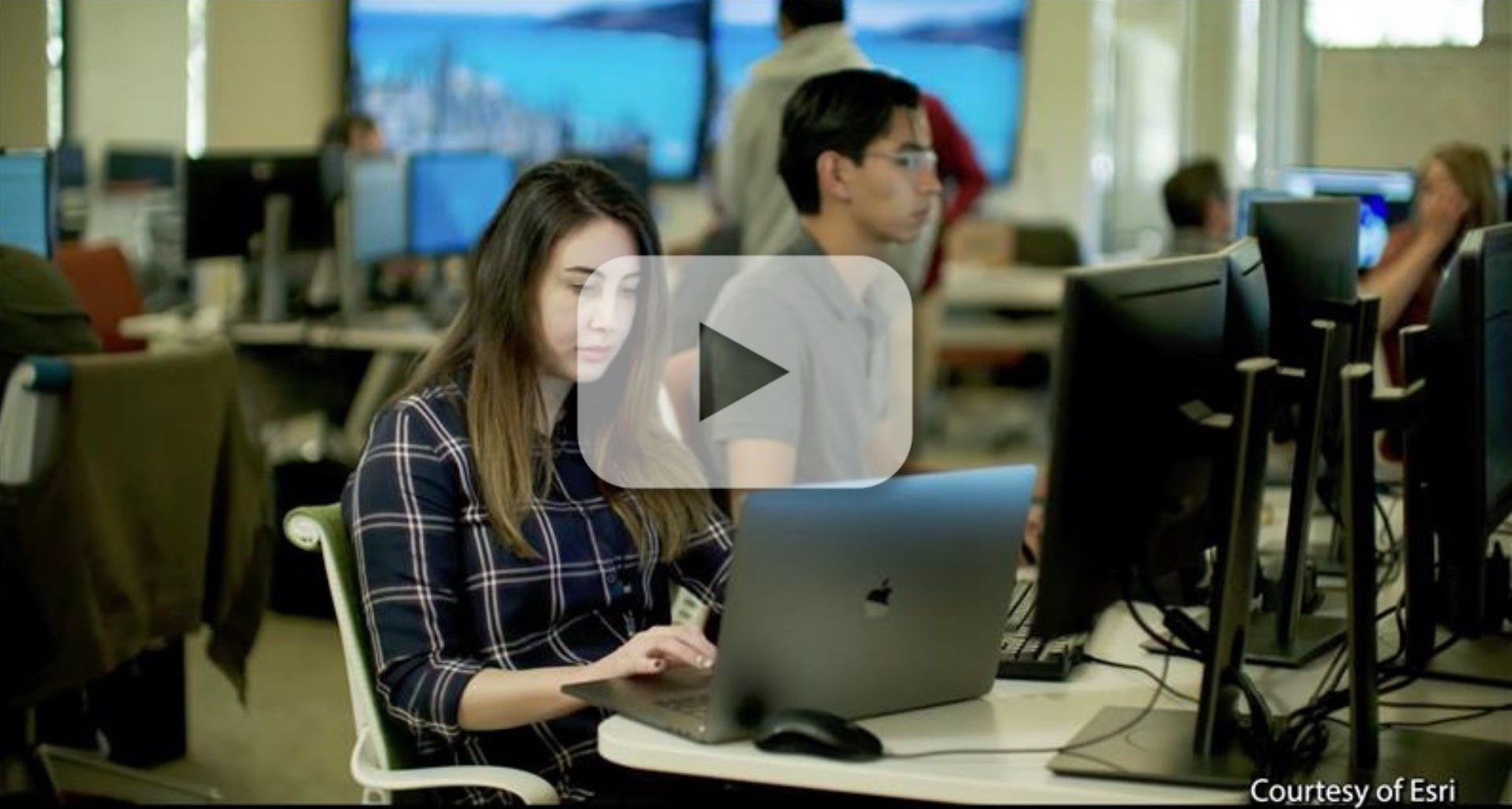 A person working on a laptop in a classroom environment with other individuals at desks using computers, courtesy of Esri.