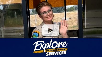 Park employee at entrance booth with "Explore San Bernardino County Services" campaign logo.