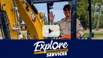A Regional Parks employee waves from a CAT excavator in a park. A blue banner below displays the Explore San Bernardino County Services logo.