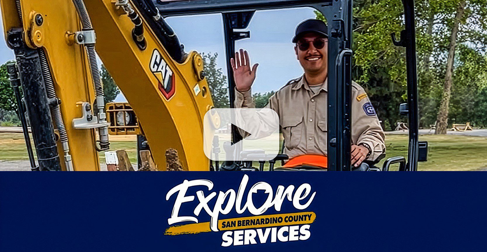 A Regional Parks employee waves from a CAT excavator in a park. A blue banner below displays the Explore San Bernardino County Services logo.