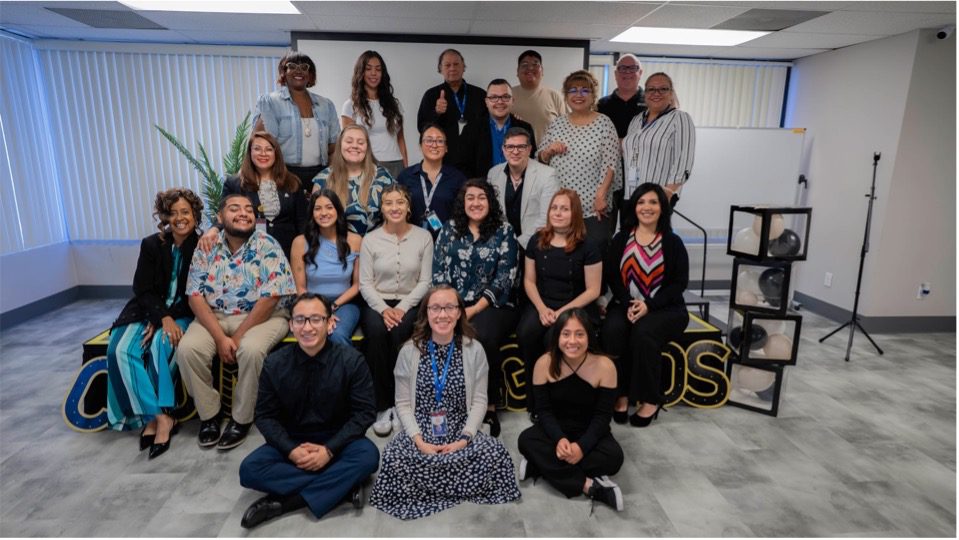 Group of people smiling for a photo in an office environment, standing and sitting in three rows, with a sign reading "Grads” in the background.