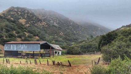 A barn in a field is surrounded by mountains.