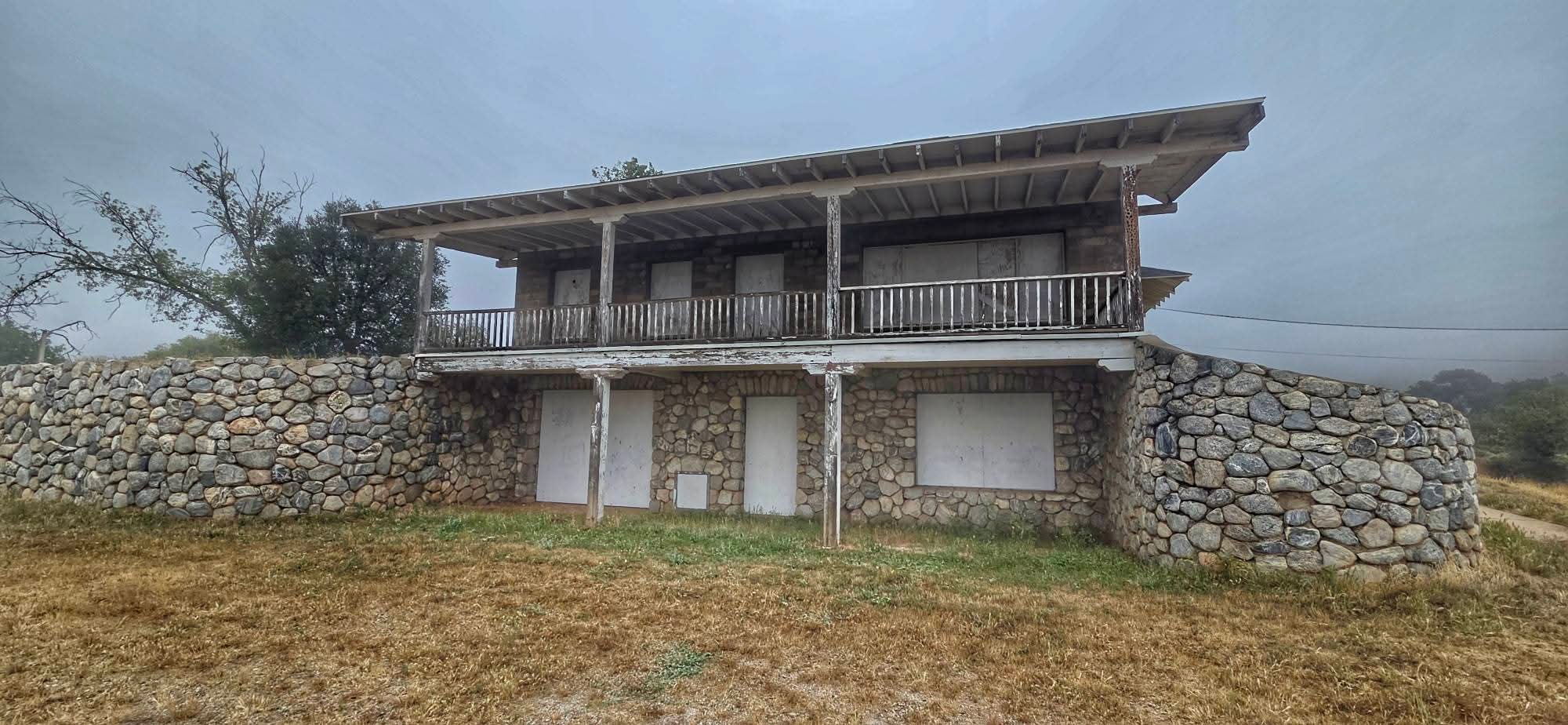 A weathered, two-story stone building with a wooden balcony stands in a grassy area. Its windows and doors appear boarded up. Trees are visible in the background under a cloudy sky.