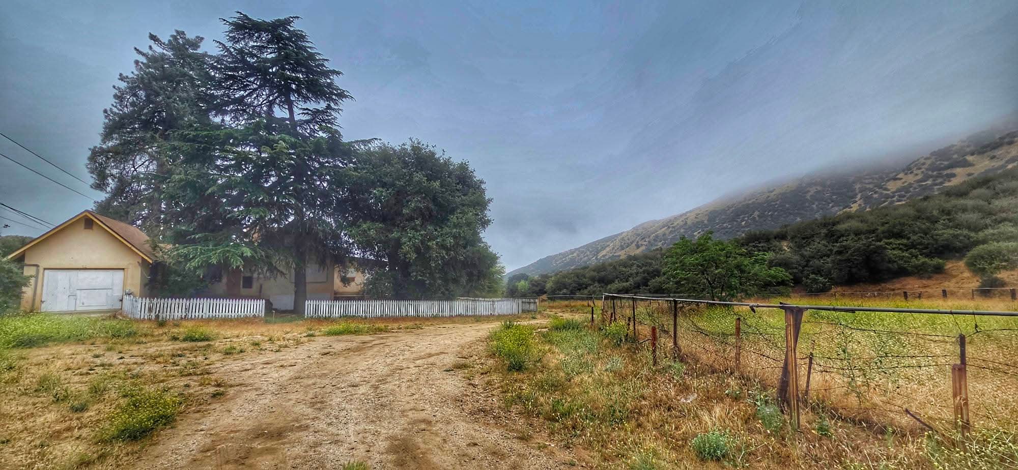 Rural landscape with a dirt road leading past a small house surrounded by trees and a white picket fence. An open field with wire fencing borders a hillside. The sky is overcast with low clouds.