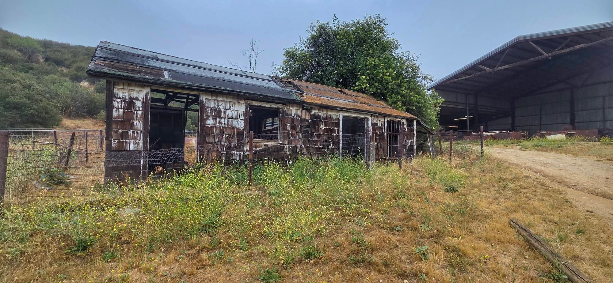 Old, weathered barn with peeling paint and damaged roof in a rural area. Surrounded by overgrown grass and a wire fence. Adjacent is a large open-sided metal structure. Forested hills in the background under a cloudy sky.