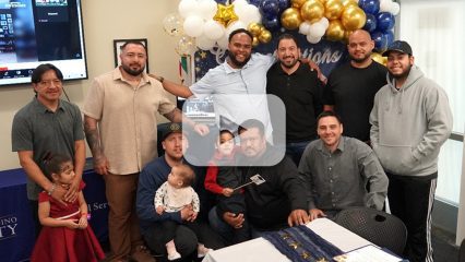 A group of nine men pose with children in front of blue, white, and gold balloons and a television.