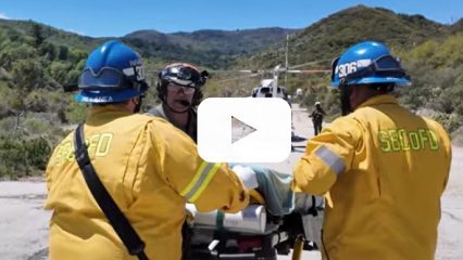 Three firemen in the national forest help a patient on a gurney to be airlifted to the hospital, with a video play button overlay indicating a video.