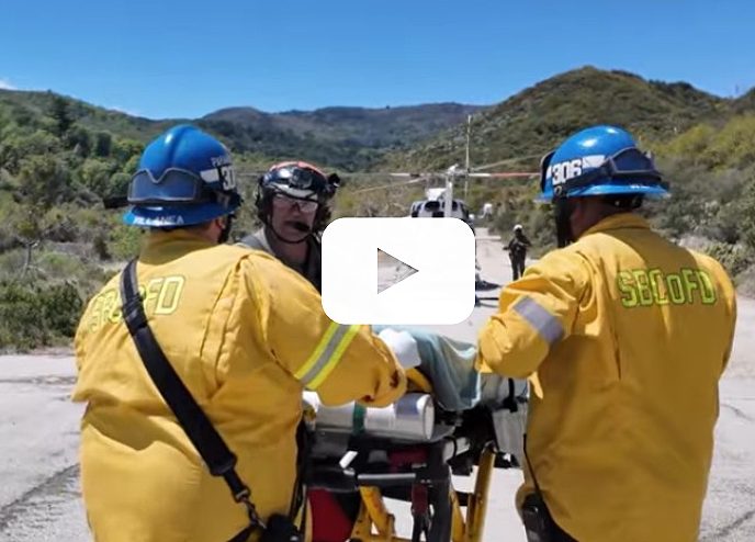 Three firemen in the national forest help a patient on a gurney to be airlifted to the hospital, with a video play button overlay indicating a video.