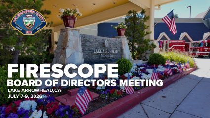 Patriotic decorations with American flags and flowers adorn a stone sign for Lake Arrowhead in front of a building. Text indicates a FIRESCOPE Board of Directors Meeting was held in Lake Arrowhead, CA, from July 7-9, 2026. A fire truck is visible in the background.