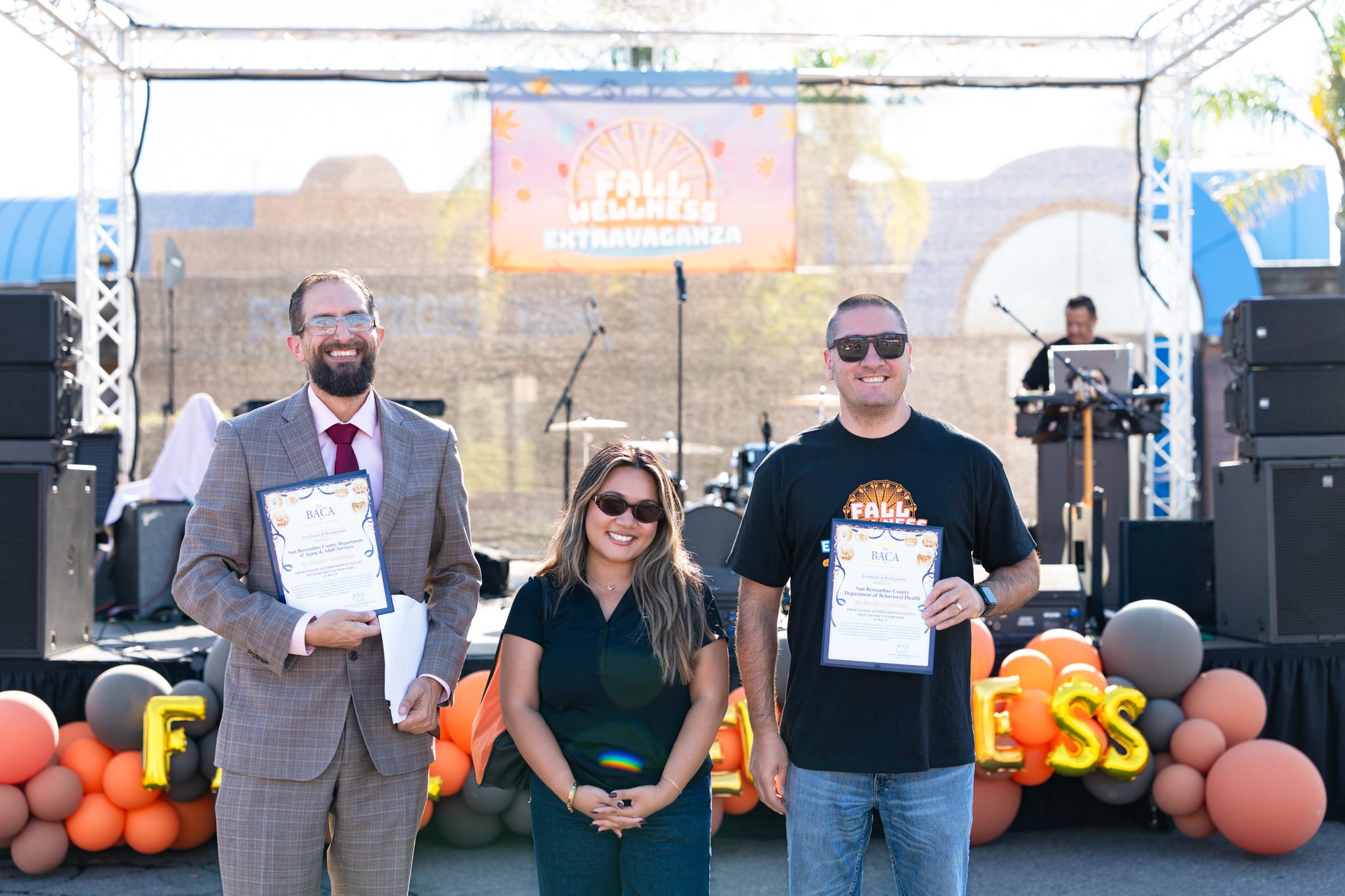 Three people pose at the Fall Wellness Extravaganza, with two holding certificates of recognition in front of the event stage.
