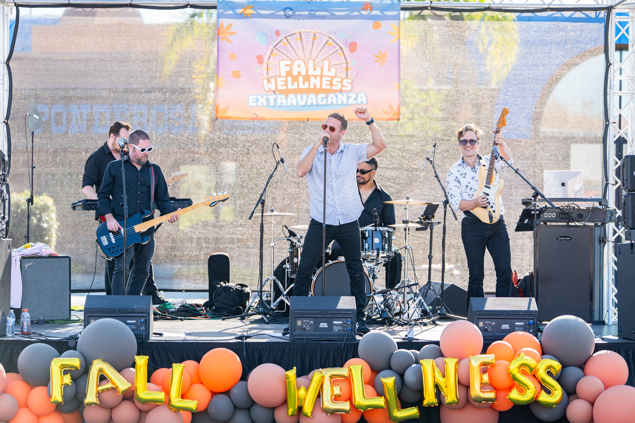A live band performs on stage at the Fall Wellness Extravaganza, surrounded by festive balloons and event decorations.
