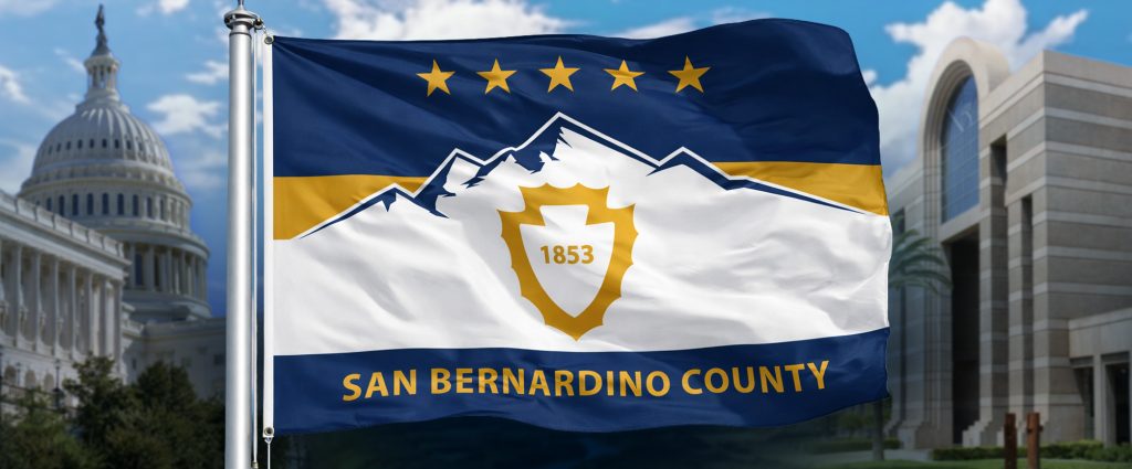 San Bernardino County flag with mountain emblem and “1853” flies before the U.S. Capitol and a civic building under blue skies.