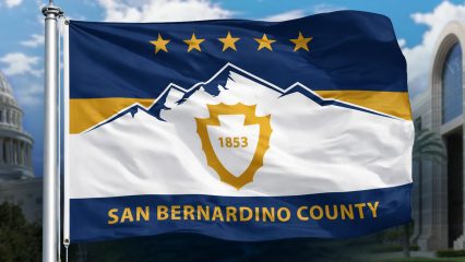 San Bernardino County flag with mountain emblem and “1853” flies before the U.S. Capitol and a civic building under blue skies.