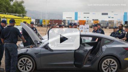 A group of San Bernardino County firefighters observes a Tesla car with the hood up, with one firefighter pointing at the engine with a play button overlay, indicating a video.
