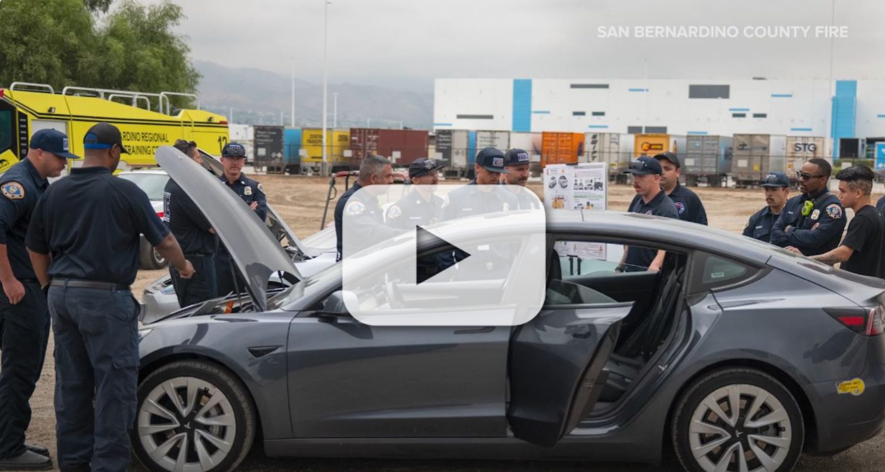 A group of San Bernardino County firefighters observes a Tesla car with the hood up, with one firefighter pointing at the engine with a play button overlay, indicating a video.