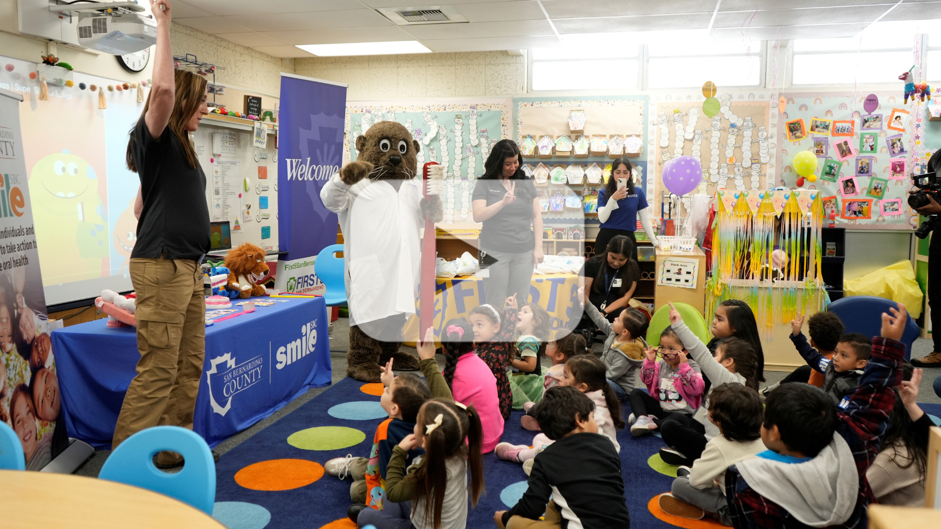 A classroom scene at Hawthorne Elementary School with preschoolers seated on a carpet for a presentation. A presenter, an otter mascot in a white lab coat holding an oversized toothbrush, and other staff members from First 5 San Bernardino are engaging the children in an activity to raise their hands. Educational materials and banners for Smile SBC and First 5 San Bernardino are displayed around the classroom. The image includes a translucent play button overlay, indicating a video.